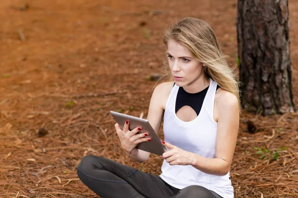 Kuru üzerinde oturan sarışın model okuma onun tablet üzerinde sopa. Gün batımında bulanık arka plan