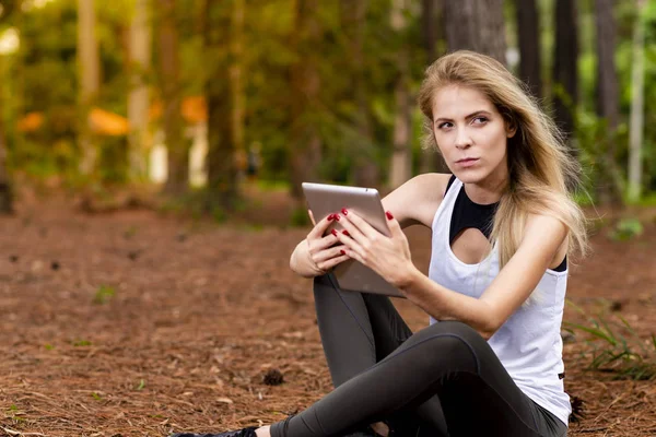 Kuru üzerinde oturan sarışın model okuma onun tablet üzerinde sopa. Gün batımında bulanık arka plan