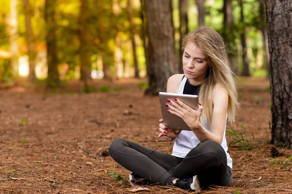 Kuru üzerinde oturan sarışın model okuma onun tablet üzerinde sopa. Gün batımında bulanık arka plan