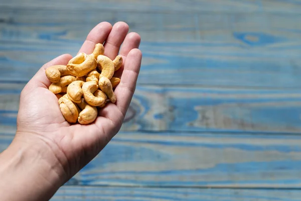Female left hand holding toasted cashew nuts over blue pine wood background.