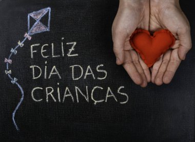 Blackboard Feliz dia das criancas (Portekizce) yazılı. Uçurtma Çizim ve kumaştan yapılmış bir kalp tutan eller. Kopyalama alanı.