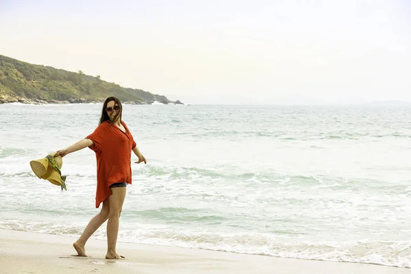 Model on deserted beach holding straw hat.