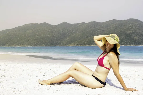 Woman in sun hat and bikini at the beach. Holding hat and laughing.