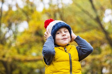 Güneşli bir sonbahar bir orman arka plan üzerinde bir ponpon ile moda sıcak örgü şapkalı küçük çocuk kameraya bakıyor