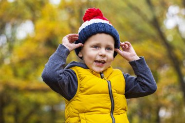 Güneşli bir sonbahar bir orman arka plan üzerinde bir ponpon ile moda sıcak örgü şapkalı küçük çocuk kameraya bakıyor
