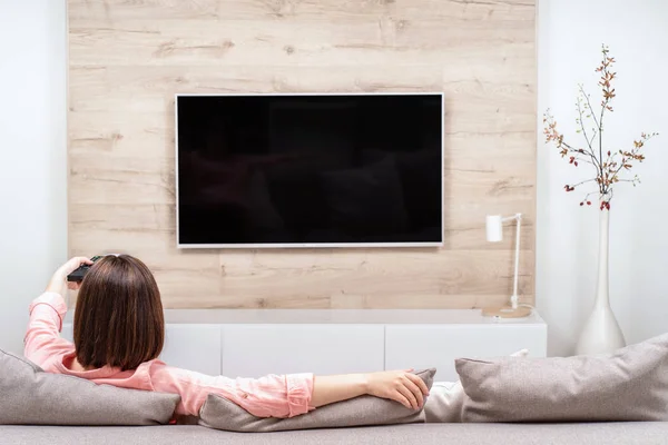 Lüks dairede dinlenen, kanepede oturan, Tv seyreden, kanal değiştiren güzel genç kadının arka görünümü.