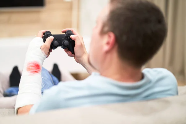 Man with broken arm in plaster cast with red blood play and holding controller in front of TV. Game addiction concept. — Stock Photo, Image