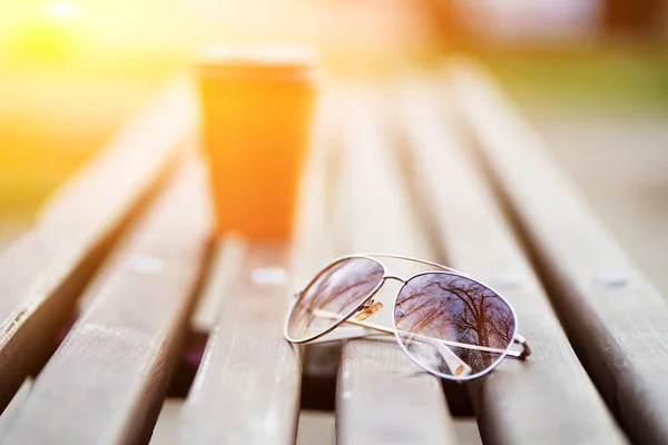 Κοντινό πλάνο φωτογραφία γυαλιών ηλίου στο ξύλινο παγκάκι στο πάρκο με θαμπή πορτοκαλί κούπα καφέ από χαρτόνι. — Φωτογραφία Αρχείου