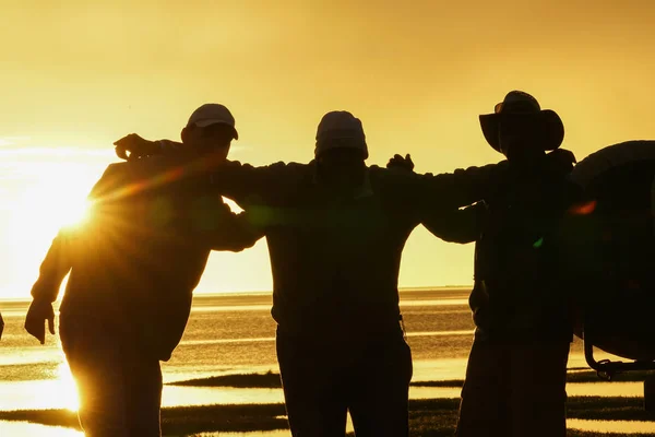 Silhouetted men back-lit by setting sun with arms around one and other