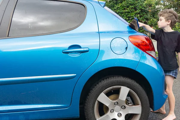 Boy earning pocket money cleaning blue compact car with hose, bucket of water and car brush