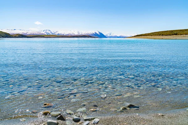 清澈干净的浅水 Tekapo 湖周围有鹅卵石底部迷人的风景和遥远的雪覆盖南阿尔卑斯山 — 图库照片