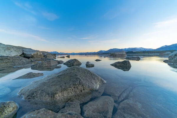 Large rocks near edge Lake Tekapo as sunsises behind distant mountains and foreshore of calm peaceful lake