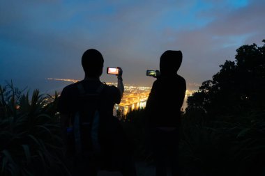 Kameralar mobil cihazlar/telefonlarda fotoğraf kentsel gece ışıkları için dönüm noktası Mount Maunganui popüler touriist hedef tepesinden kullanarak siluet grenli görüntü iki unrecognisable turist