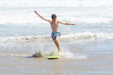 MOUNT MAUNGANUI NEW ZEALAND - FEBRUARY 10 2019: Teenager skim-boarding in shallows on beach arms outstretched for balance, clipart