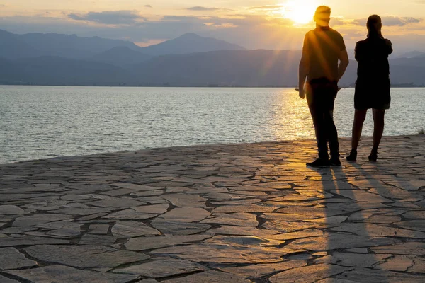 Siluetas de pareja de pie mirando al sol a través del mar . — Foto de Stock