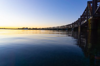Tauranga 'nın tarihi demiryolu köprüsünün sol tarafında gün doğumuyla birlikte Yeni Zelanda' nın liman ufkunda altın renkler serpiştirildi..