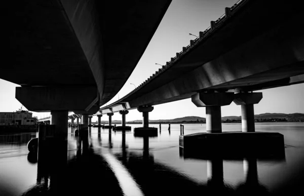 モノクロームで穏やかな水の上から下からタウランガ港橋の掃引構造線 橋はタウランガ港を渡りマウンガヌイ山へ — ストック写真