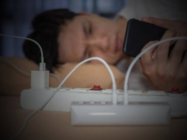 Telefonu kullanan, elektrik kablolarıyla yatağa uzanan ve telefonu şarj eden bir adam. 