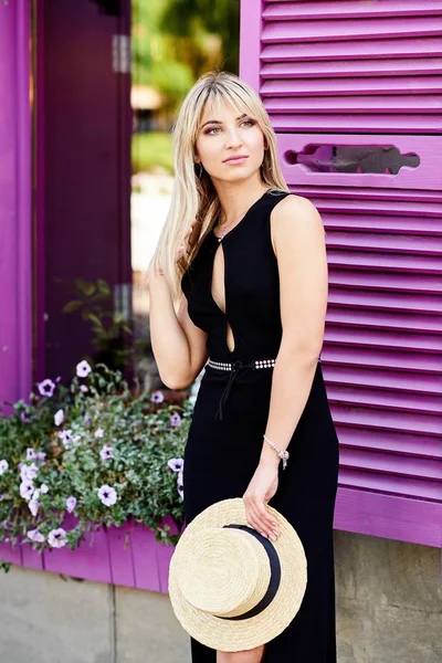 Une jeune blonde sympathique en robe noire et un chapeau de paille dans ses mains se tient sur le fond d'une fenêtre aux volets violets — Photo