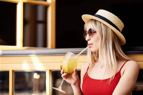 Ung ganska sympatisk blondin sitter på ett café nära ett öppet fönster med en cocktail i armarna och i en stråhatt — Stockfoto