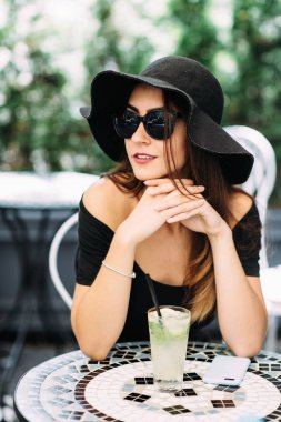 Genç güzel kız sokakta bir kafede oturan siyah şapkalı ve içecek içme soğutmalı.