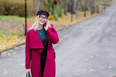 Sevimli genç kadın gözlük ve kap, kırmızı paltolu yıl sonbaharda parkta yürüyüş.