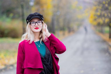 Sevimli genç kadın gözlük ve kap, kırmızı paltolu yıl sonbaharda parkta yürüyüş.