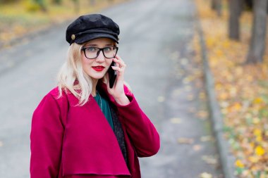 Sevimli genç kadın gözlük ve kap, kırmızı paltolu yıl sonbaharda parkta yürüyüş.