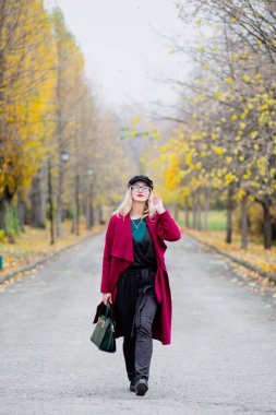 Sevimli genç kadın gözlük ve kap, kırmızı paltolu yıl sonbaharda parkta yürüyüş.