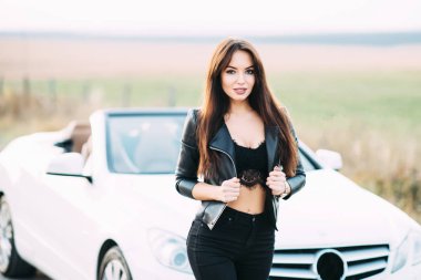 Pantolon ve beyaz cabriolet araba için bekleyen bir deri ceket giymiş kadın çok güzel.