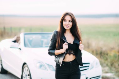 Pantolon ve beyaz cabriolet araba için bekleyen bir deri ceket giymiş kadın çok güzel.