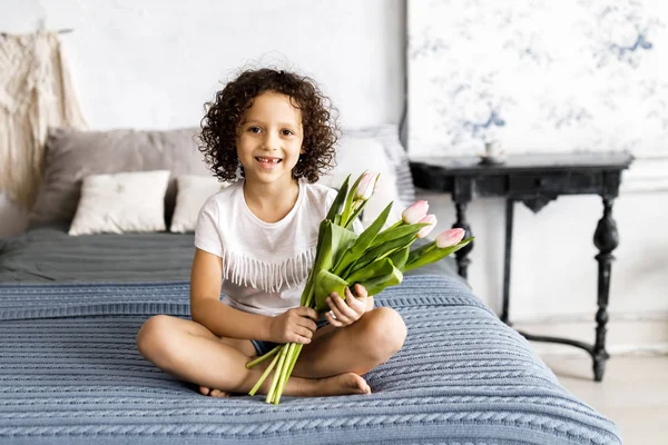A little pretty curly girl sitting on her bed with tulips in her arms