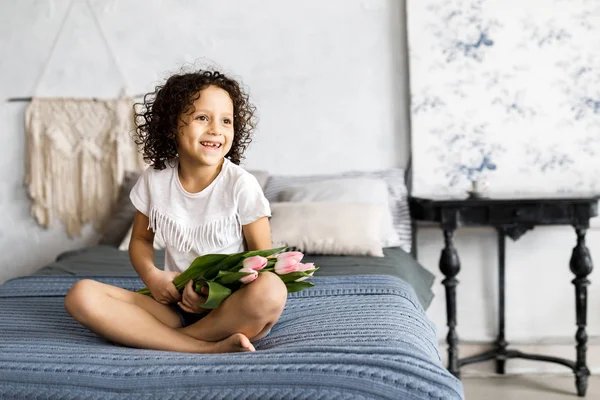 Uma menina bonita encaracolado sentado em sua cama com tulipas em seus braços — Fotografia de Stock