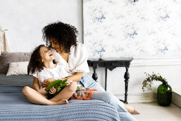 Mãe e pequena menina encaracolado bonito sentado na cama com tulipas em suas mãos — Fotografia de Stock