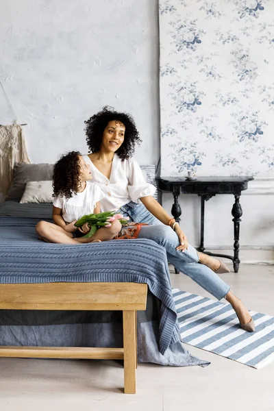 Mãe e pequena menina encaracolado bonito sentado na cama com tulipas em suas mãos — Fotografia de Stock