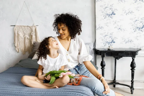 Mãe e pequena menina encaracolado bonito sentado na cama com tulipas em suas mãos — Fotografia de Stock