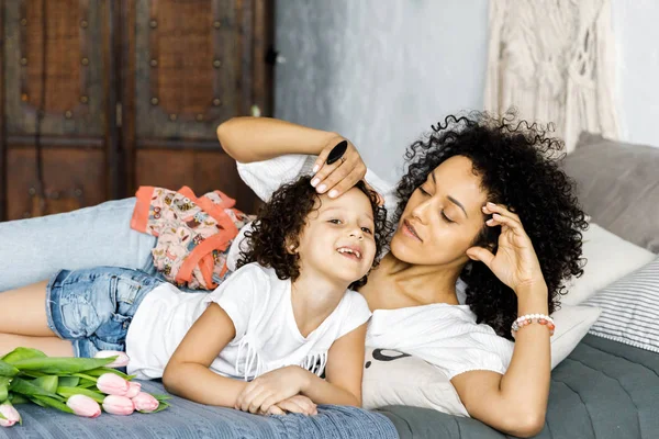 Mãe e pequena menina encaracolado bonito sentado na cama com tulipas em suas mãos — Fotografia de Stock