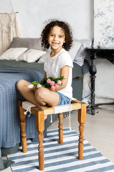 A little pretty curly girl sitting on her bed with tulips in her arms