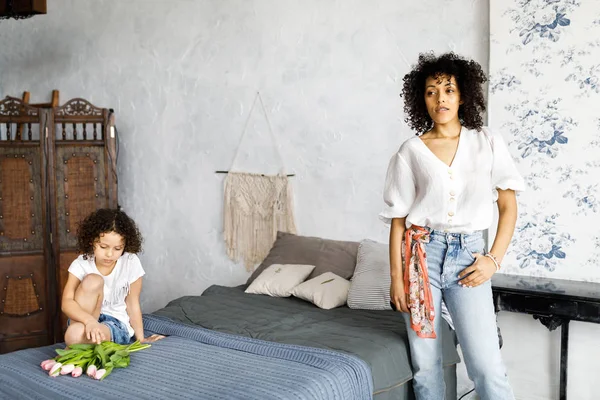 Uma mulher bonita com cabelo encaracolado está de pé no quarto ao lado da cama, e sua filha está sentada na cama e segurando uma tulipa em suas mãos. — Fotografia de Stock