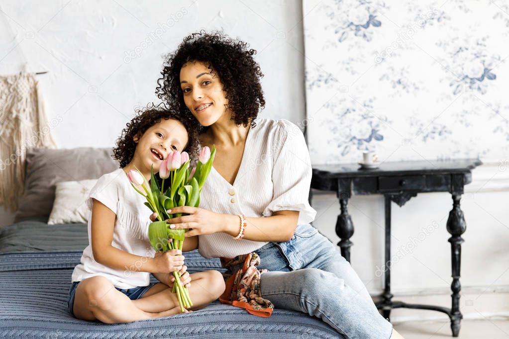 Mom and little cute curly girl sitting on bed with tulips in their hands