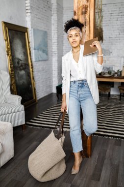 Sevimli kıvırcık kız oturma odasında duruyor ve büyük bir çanta ve tablet tutarak.