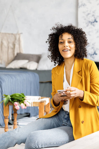 Молодая, красивая женщина с вьющимися волосами, одетая в пиджак и джинсы, сидящая на ступеньках и глядящая в телефон
