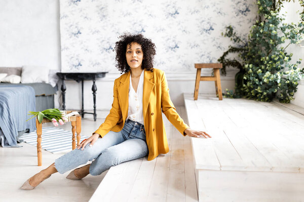 Молодая, красивая женщина с вьющимися волосами, одетая в пиджак и джинсы, сидящая на ступеньках
