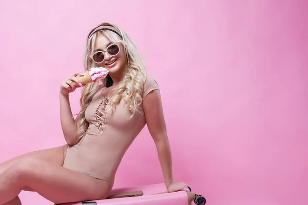Fiatal csinos lány szőke nő pózol rózsaszín háttér bőrönd és fagylalt a kezében — Stock Fotó
