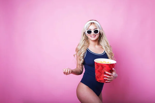Niesamowita śliczna młoda Ładna dziewczyna w okularach na różowym tle zjada popcorn i patrzy na kamerę, ubrana w niebieski strój kąpielowy. — Zdjęcie stockowe