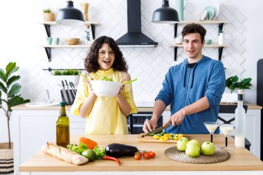 Genç, sevimli, gülümseyen bir çift mutfakta birlikte yemek pişiriyor. Gençler iyi bir mutfakta salata hazırlıyorlar.