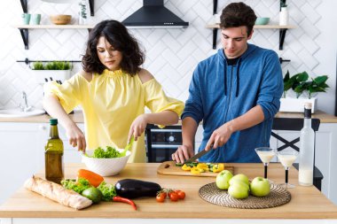 Genç, sevimli, gülümseyen bir çift mutfakta birlikte yemek pişiriyor. Gençler iyi bir mutfakta salata hazırlıyorlar.
