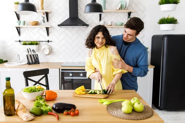 Young cute smiling couple cooking together at kitchen at home. A young woman slicing fresh vegetables on a wooden salad board. Cooking.