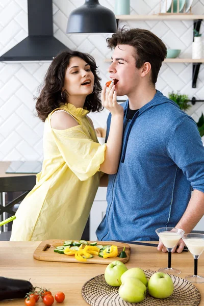 Joven linda pareja sonriente cocinar juntos en la cocina en casa. Linda chica alimenta a su novio con tomates rojos pequeños —  Fotos de Stock