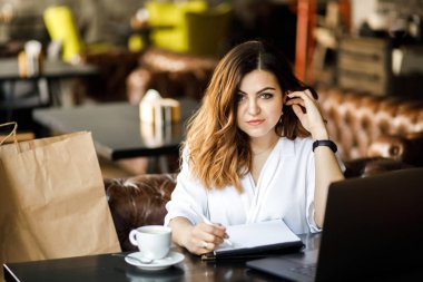 Genç, sempatik bir kadın, ince başlı bir vücut bina değil, rahat bir kafede oturur, bir bilgisayarda çalışır ve belgelere göz atar. İş giyim tarzı.